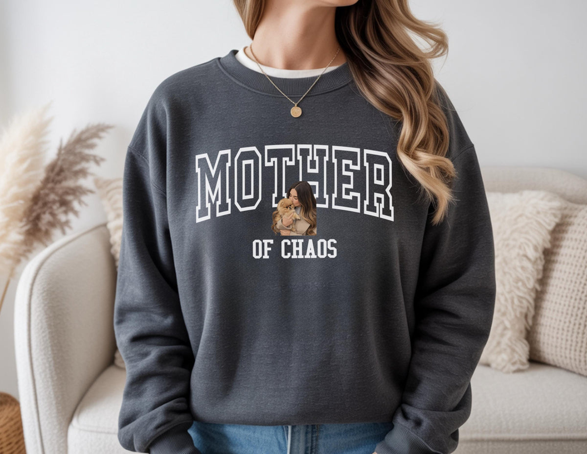 May include: Dark gray sweatshirt with the words "MOTHER of CHAOS" in white block letters. A graphic of a woman holding a child is centered below the word "MOTHER". The sweatshirt has a crew neck and long sleeves.