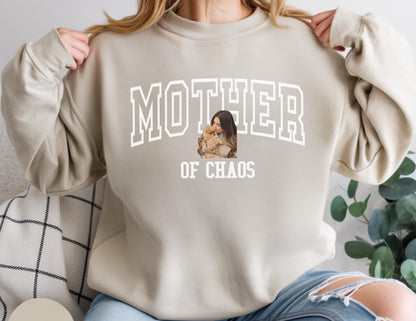May include: Beige sweatshirt with the text "MOTHER OF CHAOS" in white block letters. A graphic of a woman holding a small dog is centered under the word "MOTHER". The sweatshirt has long sleeves and a crew neck.