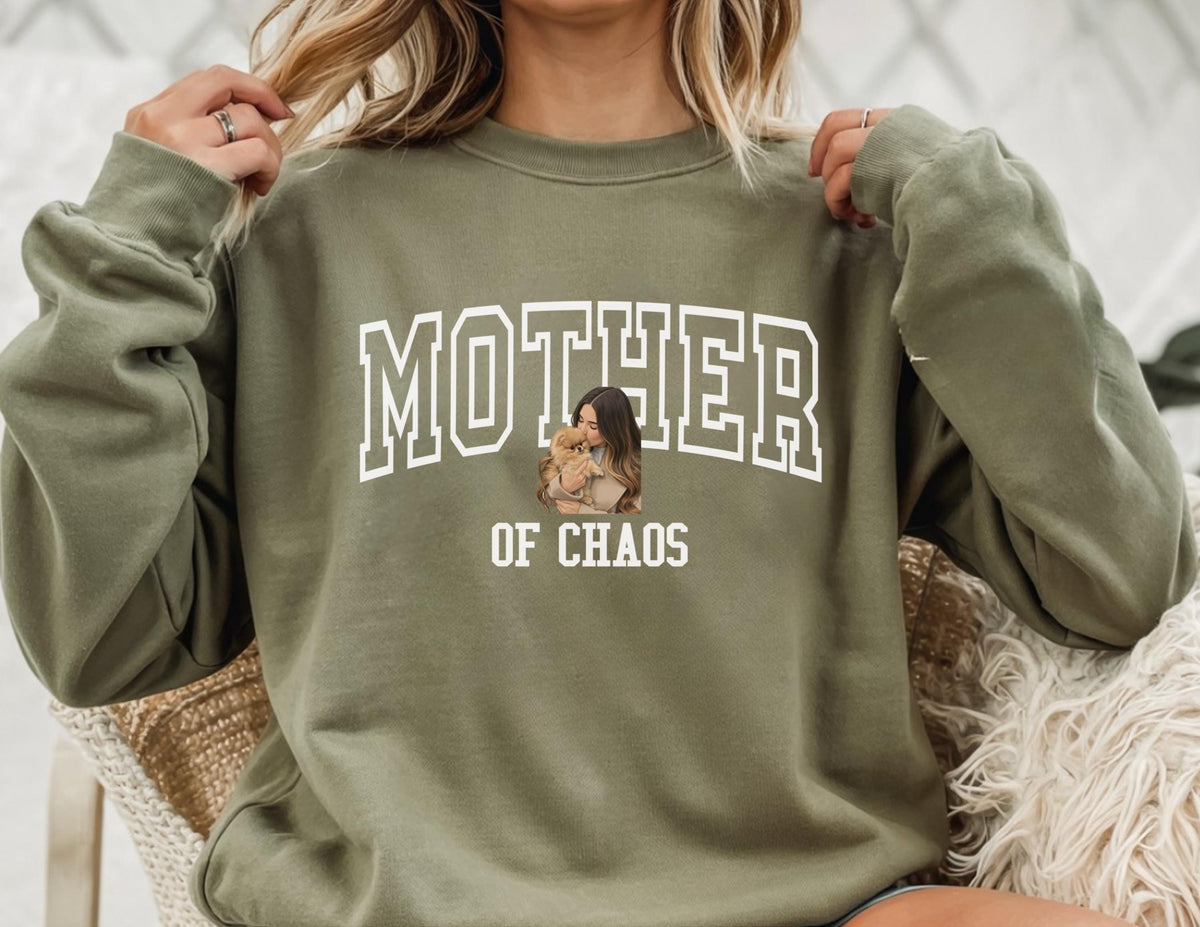 May include: Olive green sweatshirt with the text "MOTHER OF CHAOS" in white block letters. A graphic of a woman holding a child is centered below the word "MOTHER". The sweatshirt has long sleeves and a crew neck.
