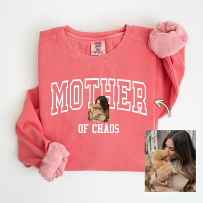 May include: Coral-colored sweatshirt with the words "MOTHER OF CHAOS" in white block letters. A small image of a woman holding a dog is centered below the word "MOTHER". The sweatshirt has long sleeves and a crew neck.