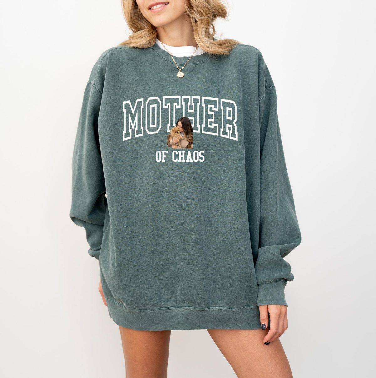 May include: A teal-colored sweatshirt with the text "MOTHER" in white block letters above "OF CHAOS". A small image of a person holding a dog is centered below the text. The sweatshirt has long sleeves and a relaxed fit.