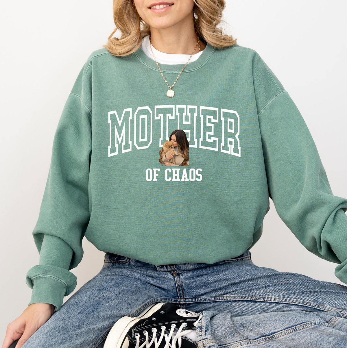 May include: Sage green sweatshirt with the text "MOTHER OF CHAOS" in white block letters. The sweatshirt features a graphic of a woman holding a small dog. The model is wearing blue jeans and black and white sneakers.