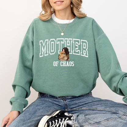 May include: Sage green sweatshirt with the text "MOTHER OF CHAOS" in white block letters. The sweatshirt features a graphic of a woman holding a small dog. The model is wearing blue jeans and black and white sneakers.