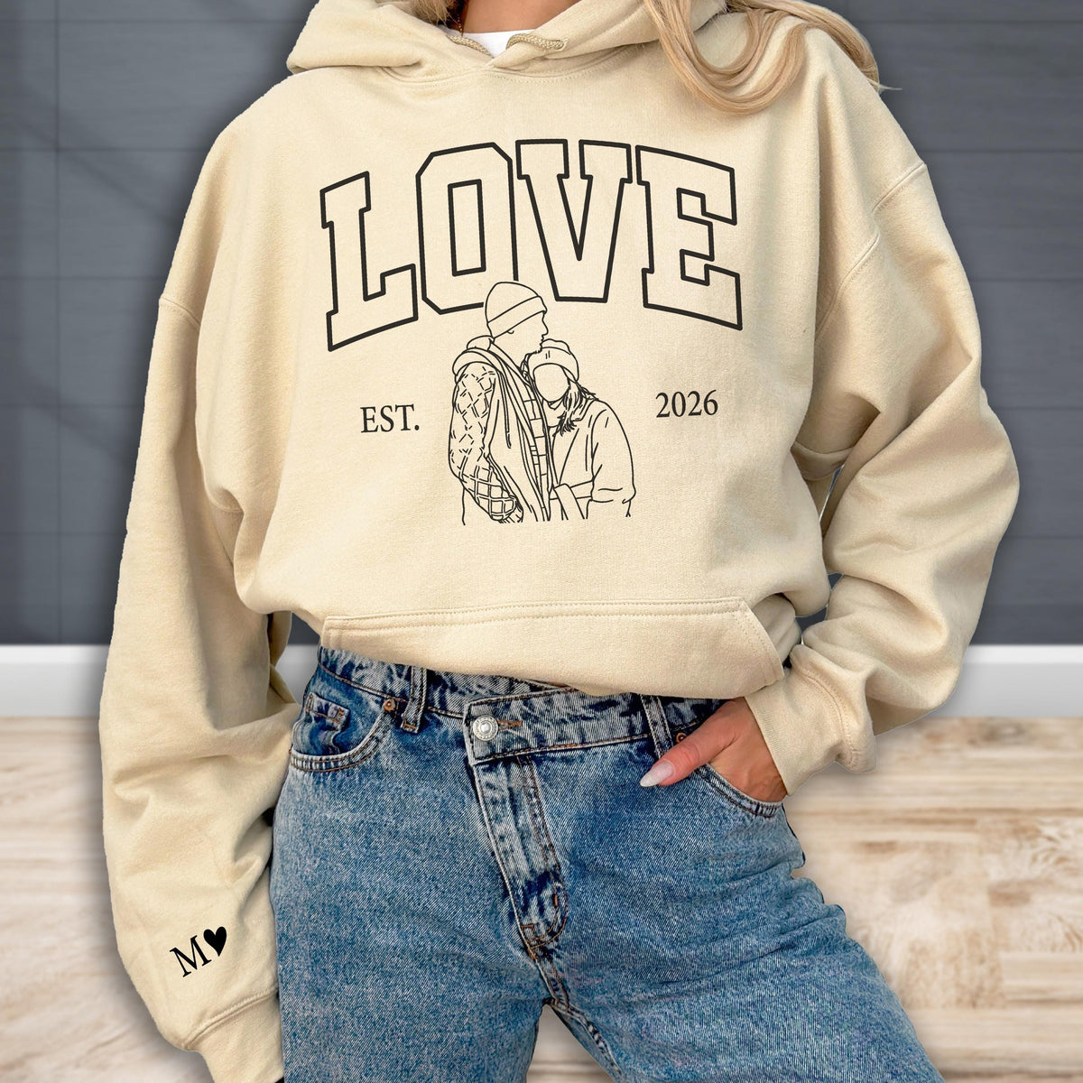 May include: A cream-colored hoodie with the word "LOVE" in bold black letters. Below the word is a black line drawing of two people embracing, with "EST. 2026" printed on either side. The left sleeve has a black "M" with a heart.