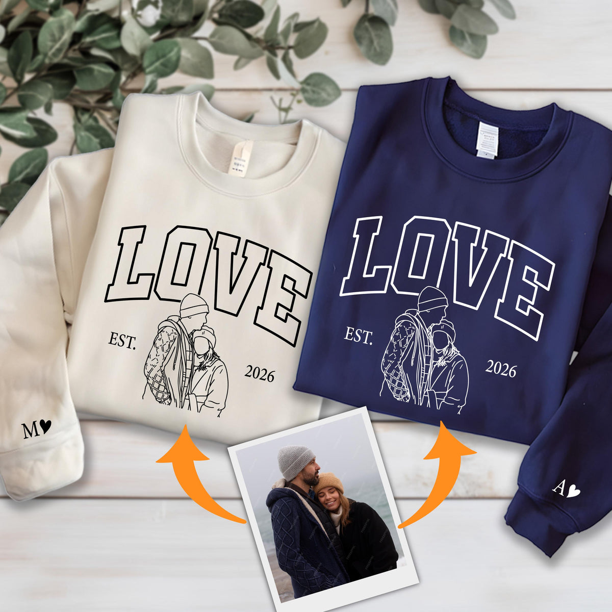 May include: Two crewneck sweatshirts, one cream and one navy blue, with the word "LOVE" in bold white letters. Below the word is a line drawing of a couple and the text "EST. 2026". The cream sweatshirt has an "M" and a heart on the sleeve, and the navy one has an "A" and a heart.