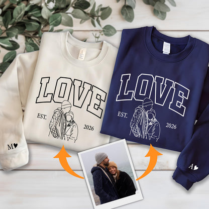 May include: Two crewneck sweatshirts, one cream and one navy blue, with the word "LOVE" in bold white letters. Below the word is a line drawing of a couple and the text "EST. 2026". The cream sweatshirt has an "M" and a heart on the sleeve, and the navy one has an "A" and a heart.