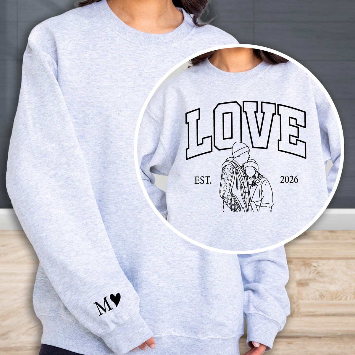May include: A light gray sweatshirt with the word "LOVE" in a collegiate font, with a line drawing of a couple and the year "2026". The left sleeve has the initials "M" and a heart. The sweatshirt is a casual style.