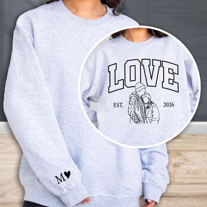 May include: A light gray sweatshirt with the word "LOVE" in a collegiate font, with a line drawing of a couple and the year "2026". The left sleeve has the initials "M" and a heart. The sweatshirt is a casual style.