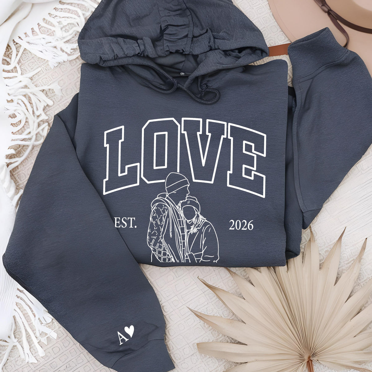 May include: A dark gray hoodie with the word "LOVE" in white block letters. Below the word is a line drawing of two people and the text "EST. 2026". The left sleeve has a white heart and the initials "A V".