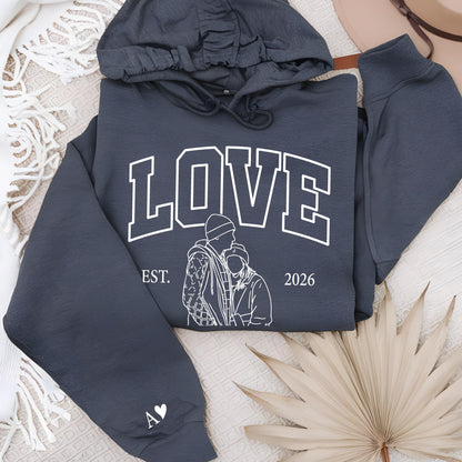 May include: A dark gray hoodie with the word "LOVE" in white block letters. Below the word is a line drawing of two people and the text "EST. 2026". The left sleeve has a white heart and the initials "A V".