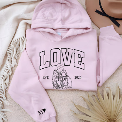 May include: A light pink hoodie with the word "LOVE" in black block letters. Below the word "LOVE" is the text "EST. 2026" and a black and white illustration of a couple. The left sleeve has a black heart with the letter "M".
