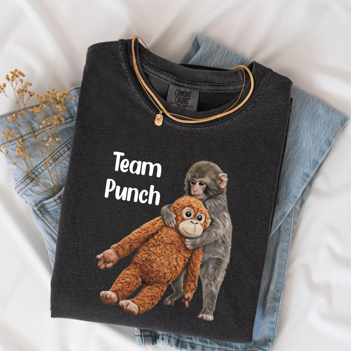 May include: Dark gray t-shirt with the text "Team Punch" and a graphic of two monkeys hugging. One monkey is brown, the other is gray. A gold necklace is draped over the shirt, which is folded on top of blue jeans.