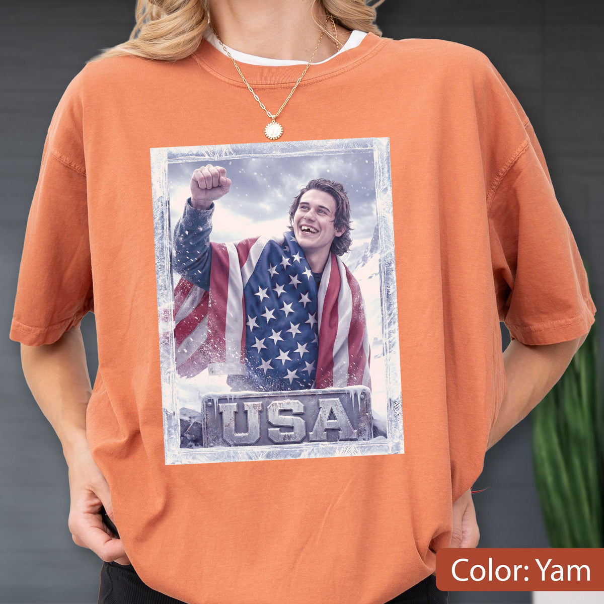 May include: Yam-colored t-shirt featuring a framed image of a person draped in an American flag, with the word "USA" at the bottom. The person is smiling and has their fist raised. The shirt has short sleeves.