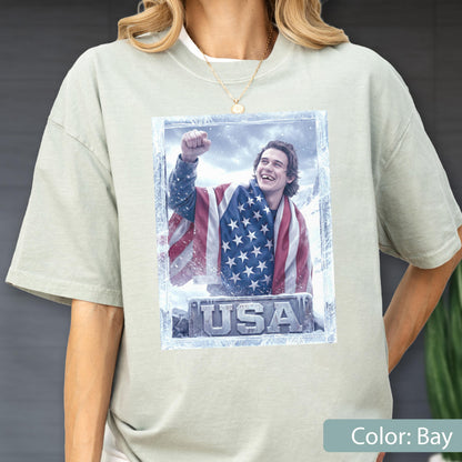 May include: Bay-colored t-shirt featuring a graphic of a person wrapped in an American flag, with the word "USA" at the bottom. The graphic is framed with a frosty border. The person is smiling and raising a fist.