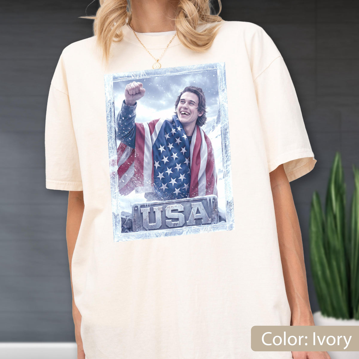 May include: Ivory-colored t-shirt featuring a graphic of a person wrapped in an American flag, with the word "USA" at the bottom. The graphic is framed in a light blue border. The shirt has short sleeves and a relaxed fit.