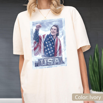 May include: Ivory-colored t-shirt featuring a graphic of a person wrapped in an American flag, with the word "USA" at the bottom. The graphic is framed in a light blue border. The shirt has short sleeves and a relaxed fit.