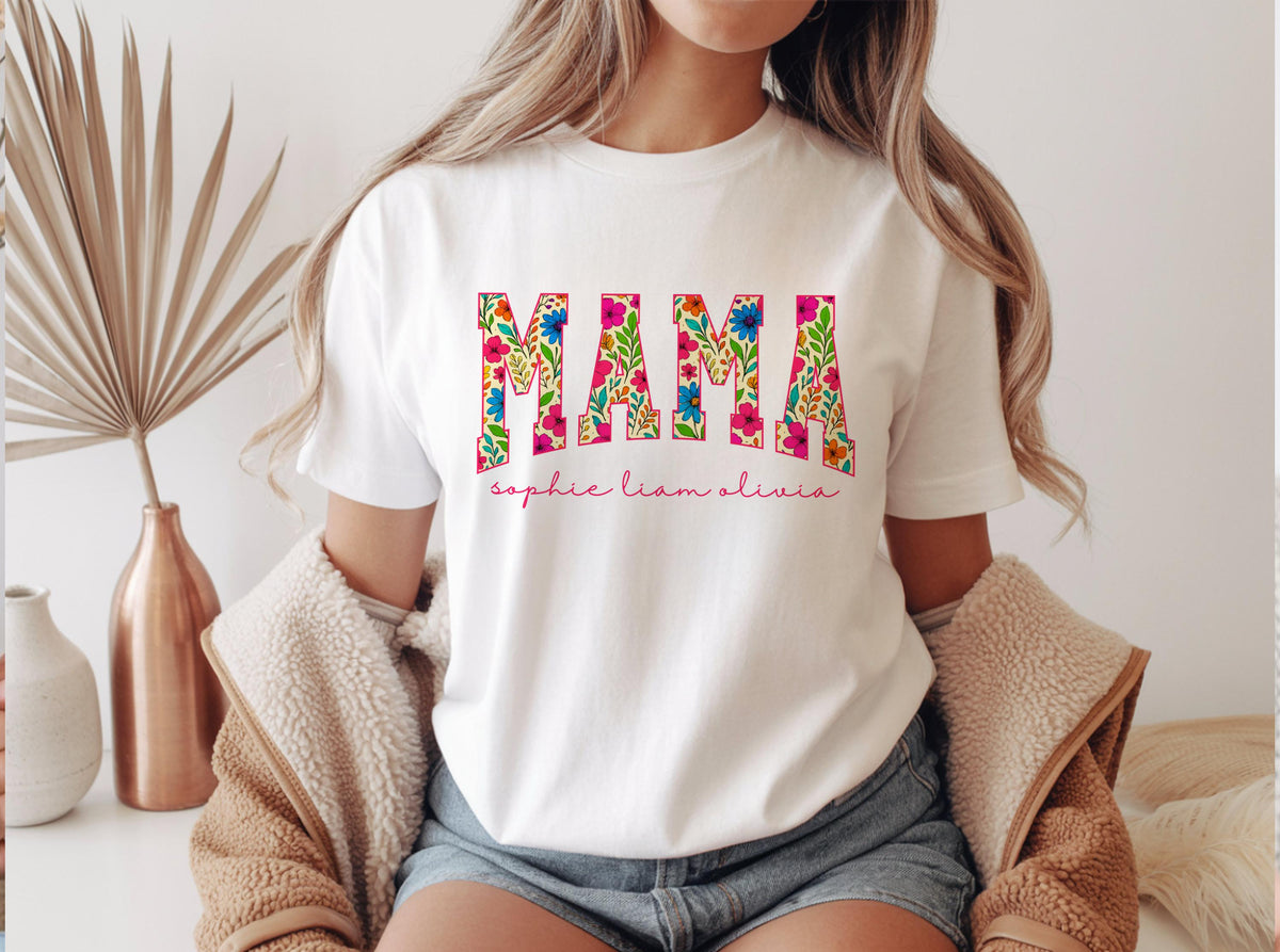 May include: White t-shirt with the word "MAMA" in large, floral-patterned letters. Below the word "MAMA" are the names "sophie liam olivia" in a cursive font. The shirt is worn by a person.