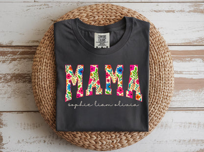 May include: Dark gray t-shirt with the word "MAMA" in a floral design. Below the word "MAMA" are the names "sophie liam olivia" in a cursive font. The shirt is displayed on a woven, circular mat.