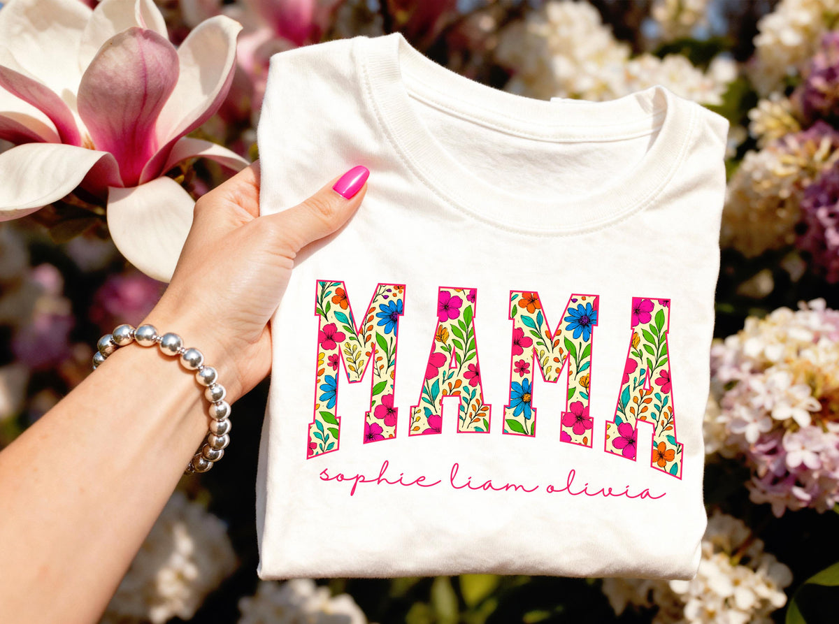 May include: White t-shirt with the word "MAMA" in a floral design. The letters are filled with colorful flowers and green leaves. Below the word "MAMA" is the text "sophie liam olivia" in a cursive font.