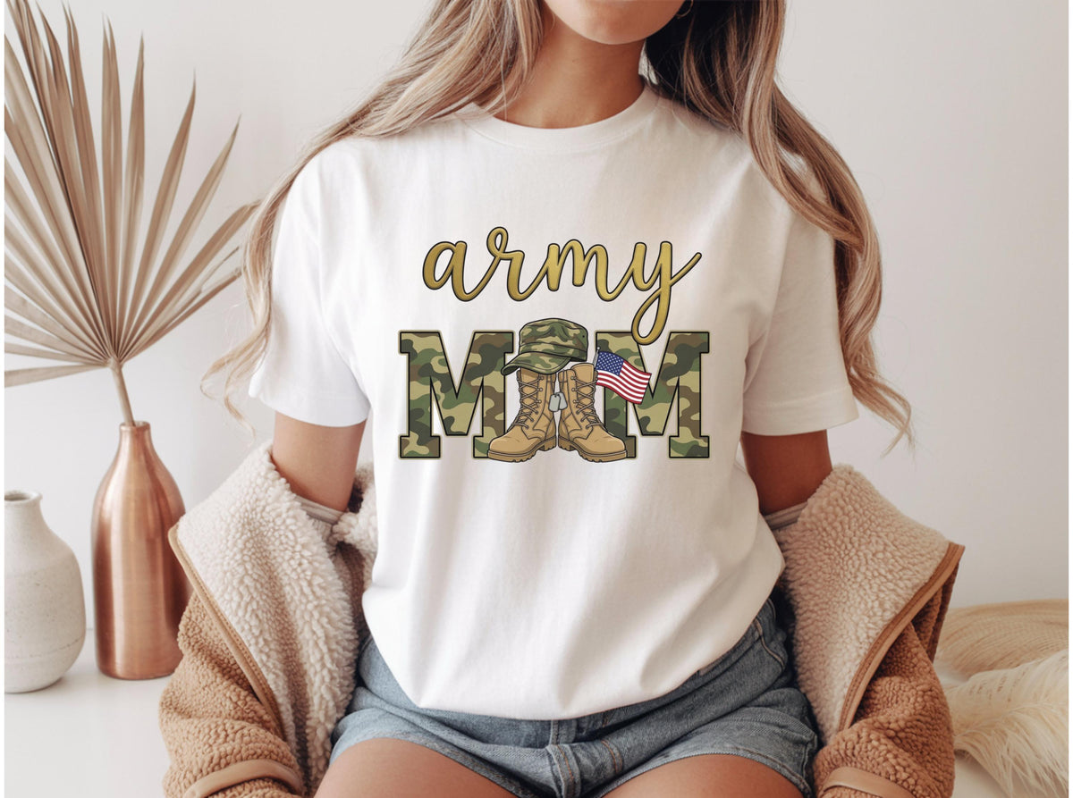 May include: White t-shirt with the text "army MOM" in gold script and camouflage block letters. The design includes a military hat, boots, and an American flag. The shirt is a casual style.