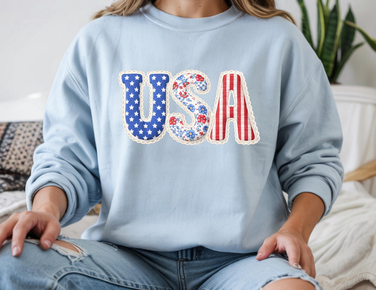 May include: A light blue sweatshirt with the word "USA" in a patriotic design. The "U" is filled with white stars on a blue background, the "S" has a floral pattern, and the "A" has red and white stripes. The letters are outlined with a white lace trim.