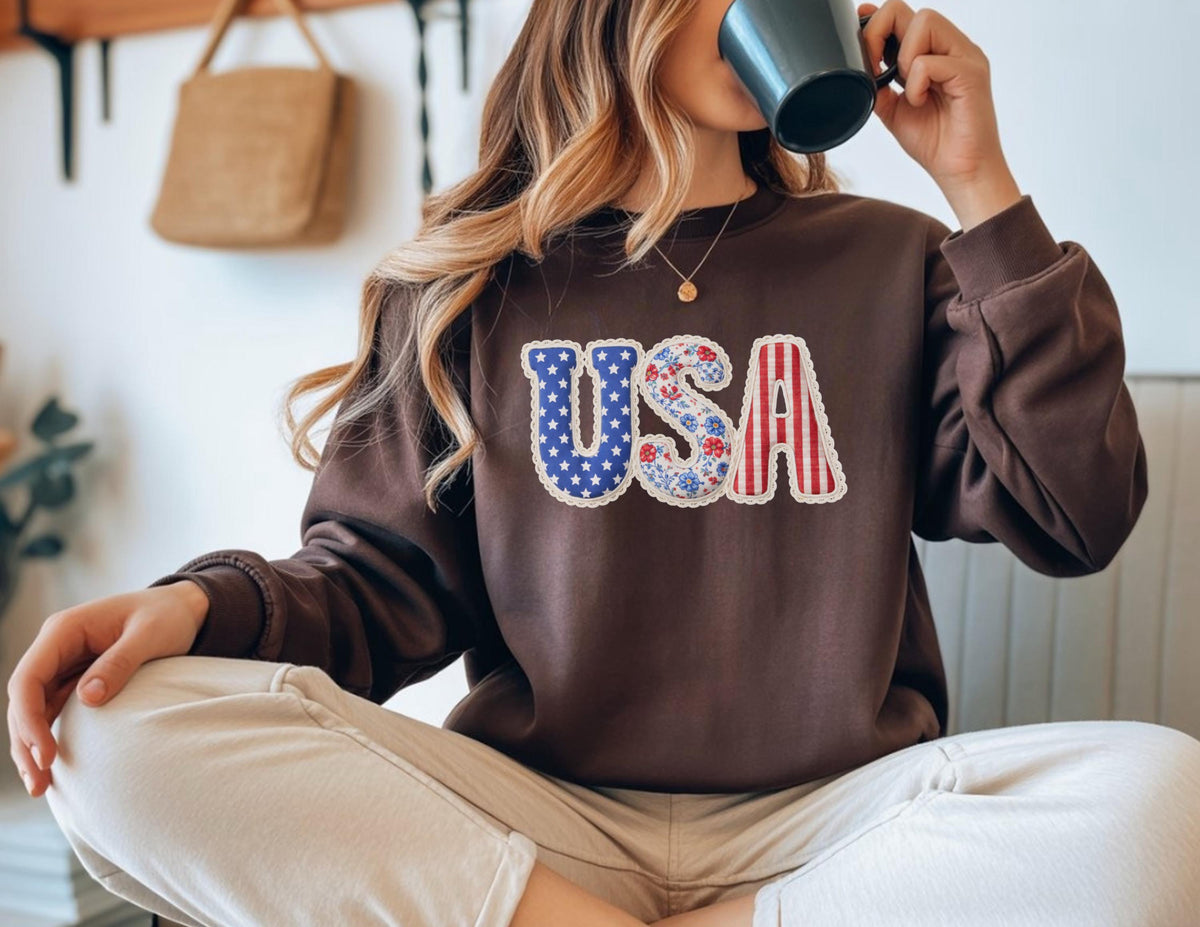 May include: A brown sweatshirt with the word "USA" in a patriotic design. The "U" is blue with white stars, the "S" has a floral pattern, and the "A" has red and white stripes. The sweatshirt is a solid brown color.