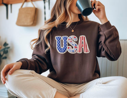 May include: A brown sweatshirt with the word "USA" in a patriotic design. The "U" is blue with white stars, the "S" has a floral pattern, and the "A" has red and white stripes. The sweatshirt is a solid brown color.