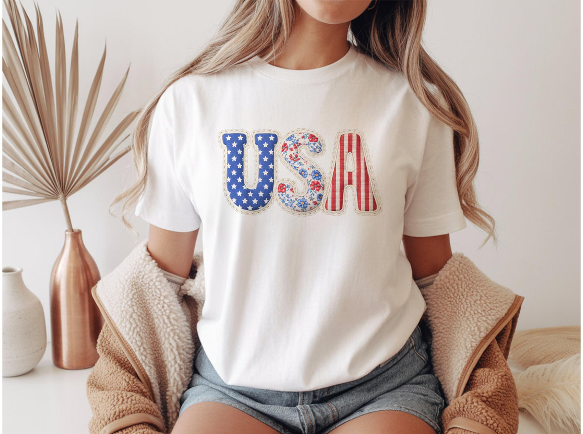 May include: White t-shirt with the word "USA" in patriotic colors. The "U" is blue with white stars, the "S" has a floral pattern, and the "A" is red and white striped. A casual, everyday tee.