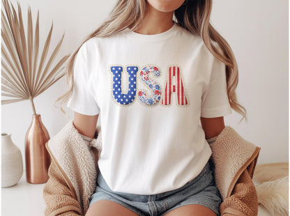 May include: White t-shirt with the word "USA" in patriotic colors. The "U" is blue with white stars, the "S" has a floral pattern, and the "A" is red and white striped. A casual, everyday tee.