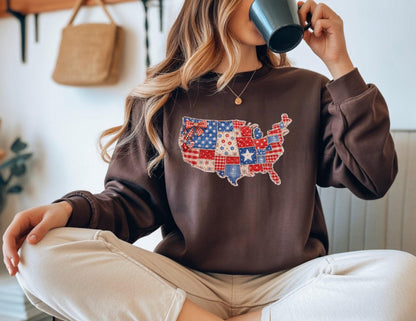 May include: A brown sweatshirt featuring a patchwork design of the United States map in red, white, and blue. The design incorporates stars, stripes, and various patterns. The sweatshirt has a crew neck and long sleeves.
