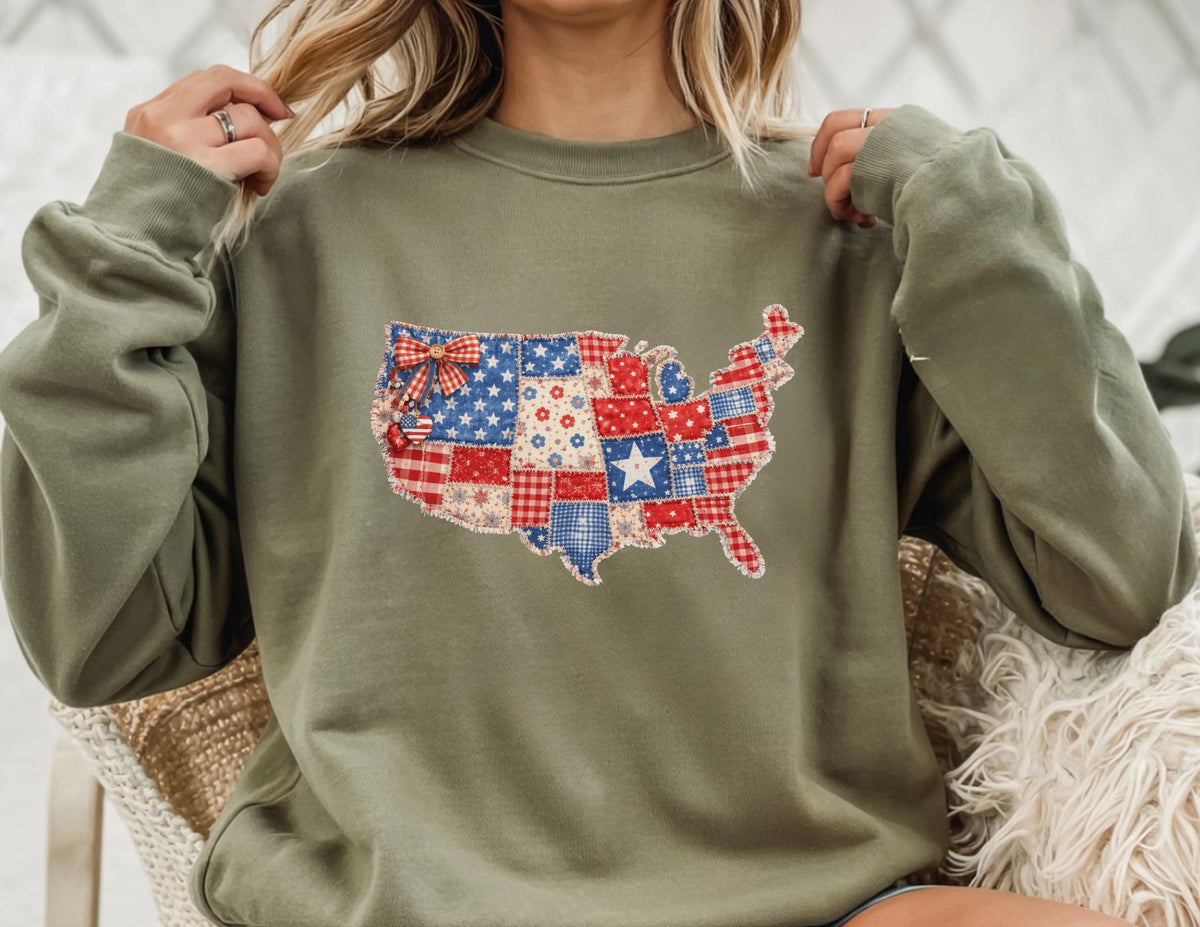 May include: Olive green sweatshirt featuring a patchwork design of the United States map. The map is composed of red, white, and blue fabric squares, stars, and a bow. The sweatshirt has long sleeves and a crew neck.