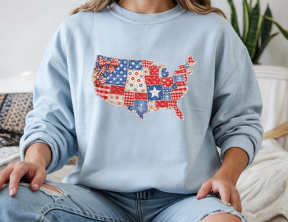 May include: A light blue sweatshirt featuring a patchwork design of the United States map. The map is composed of red, white, and blue fabrics with star and bow accents. The sweatshirt has long sleeves and a crew neck.