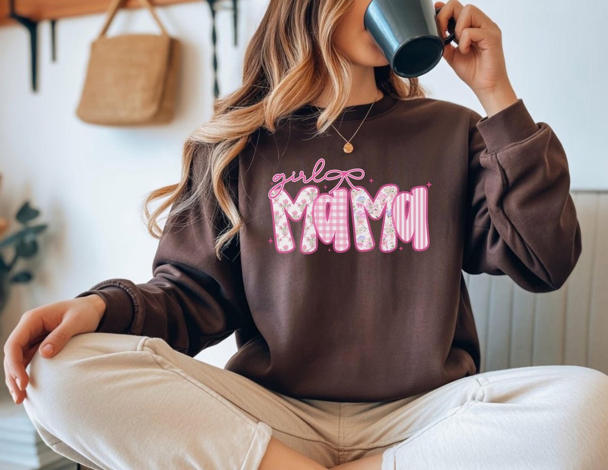 May include: A brown sweatshirt with the text "girl mama" in pink, featuring a bow and patterned letters. The letters have a gingham, floral, and striped design. The sweatshirt has long sleeves and a crew neck.