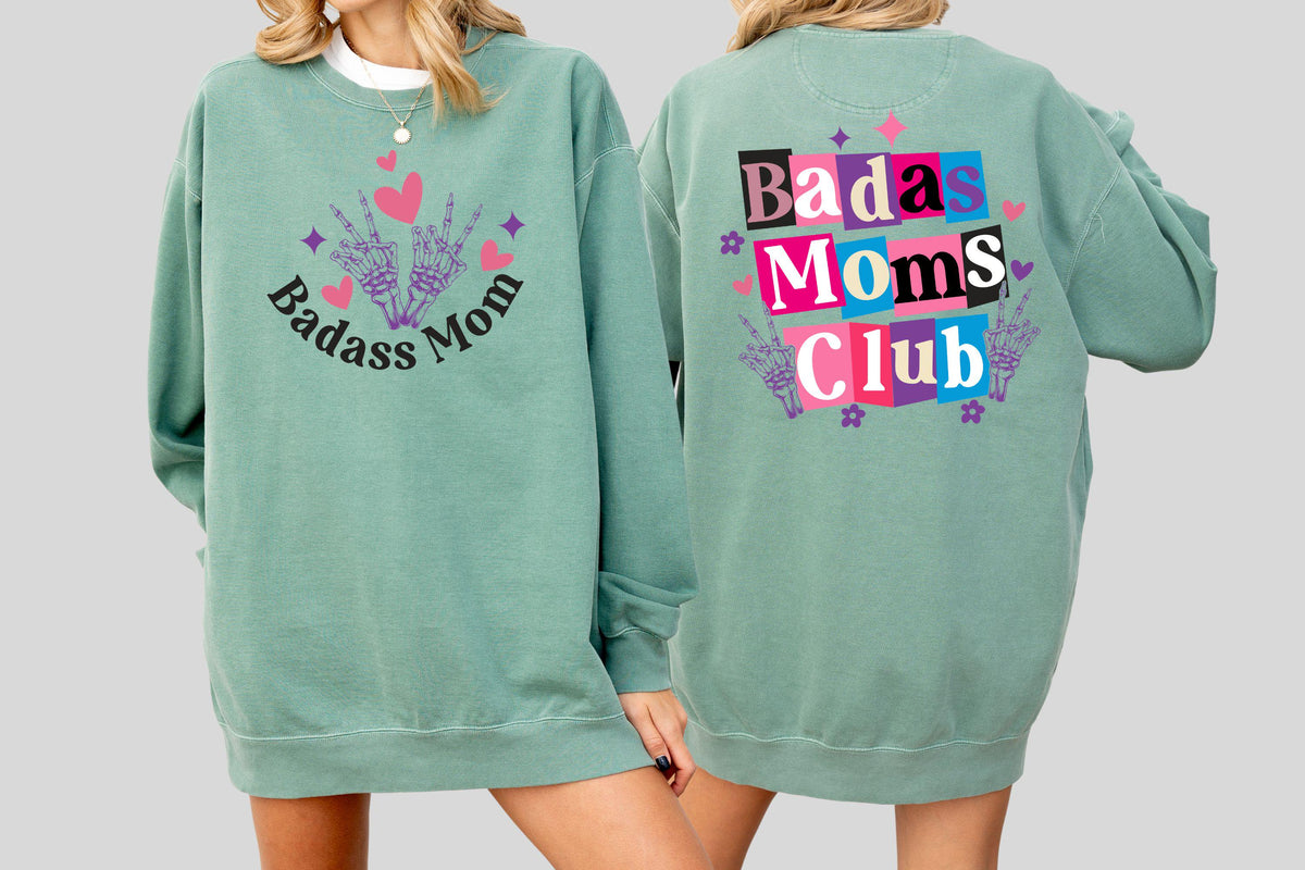 May include: A sage green sweatshirt with the words "Badass Mom" and a hand gesture graphic on the front. The back of the sweatshirt features the text "Badass Moms Club" in a colorful block design with additional graphics.