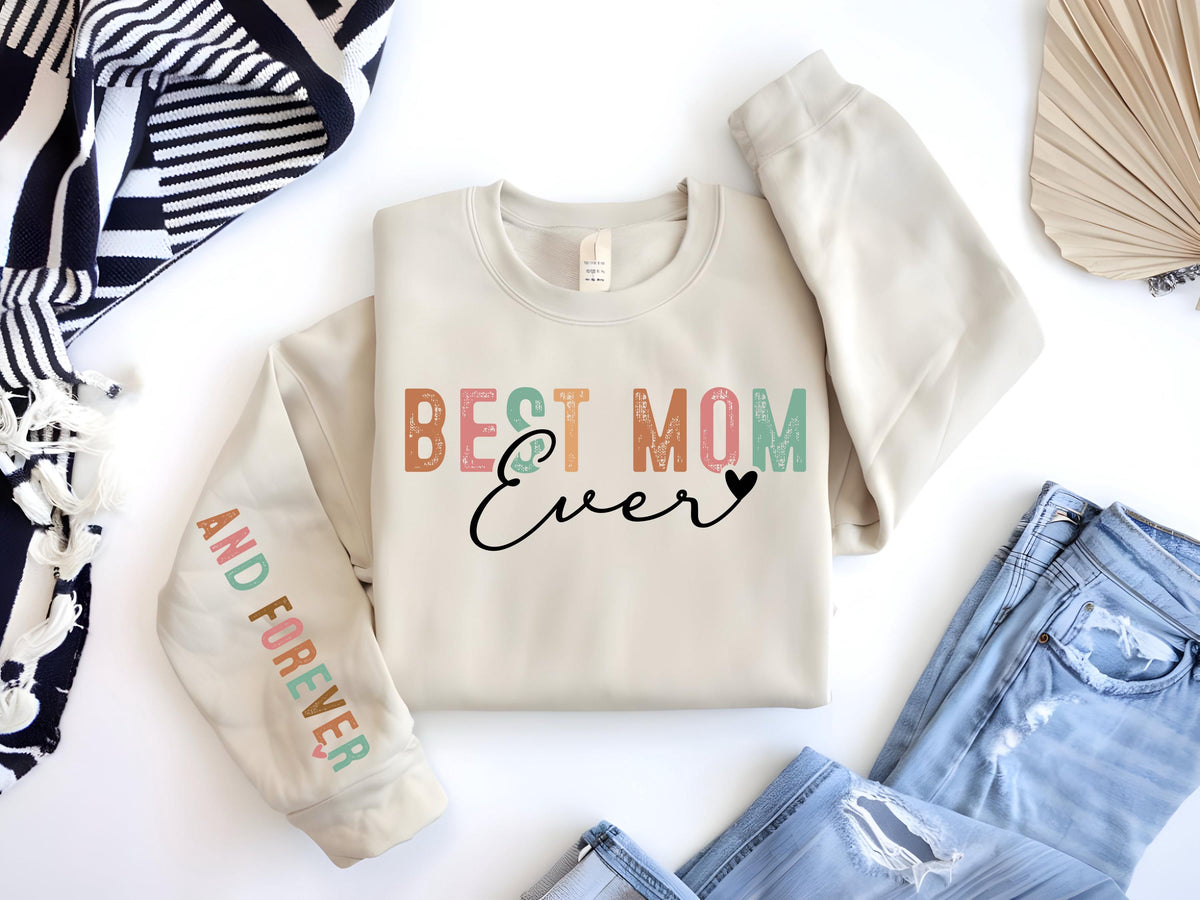 May include: Cream-colored sweatshirt with the text "BEST MOM Ever" in colorful letters and a black heart. The sleeve reads "AND FOREVER" in pastel colors. The sweatshirt is paired with blue jeans.