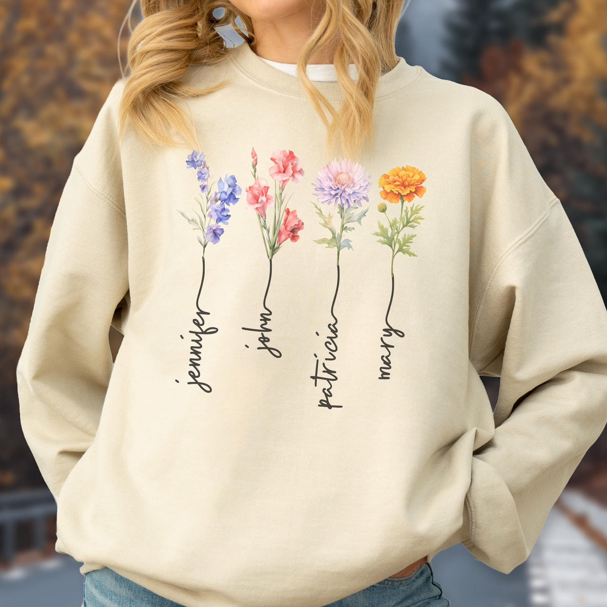May include: Beige sweatshirt featuring a floral design with the names "Jennifer", "John", "Patricia", and "Mary" in a script font. The flowers are in shades of purple, pink, and orange.