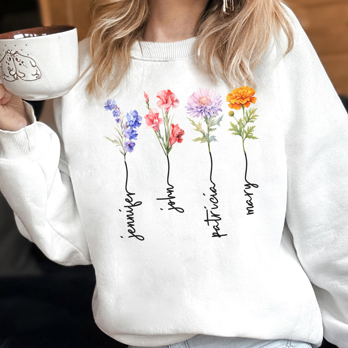 May include: White sweatshirt featuring four colorful floral designs with names Jennifer, John, Patricia, and Mary in a script font. Each flower is a different color, including blue, red, pink, and orange. The sweatshirt is a casual, personalized item.