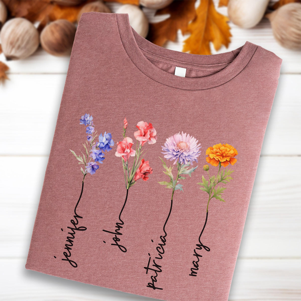 May include: A mauve-colored t-shirt featuring a floral design with the names "Jennifer", "John", "Patricia", and "Mary" printed below the flowers. The flowers are in shades of blue, pink, purple, and orange.