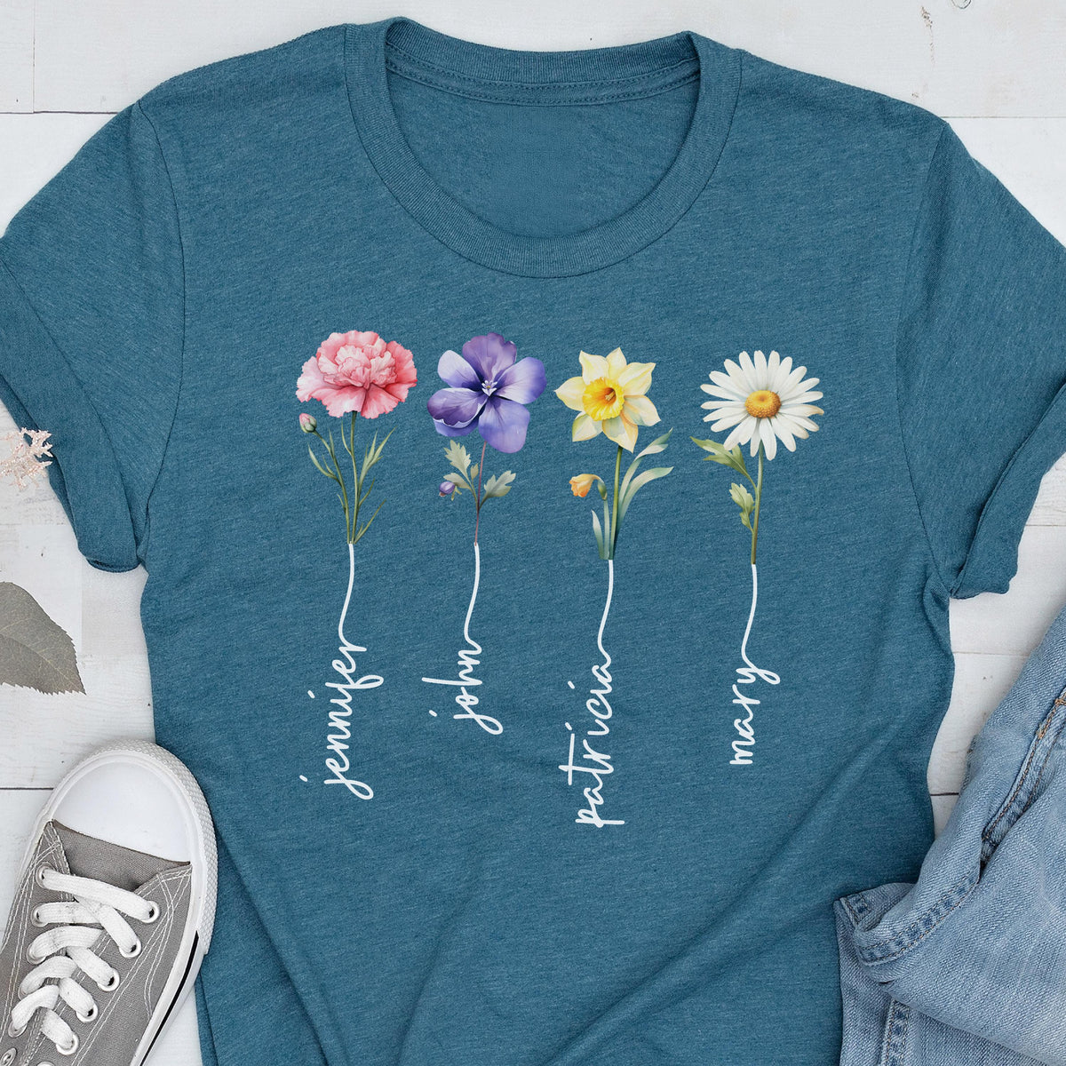 May include: Teal t-shirt featuring a floral design with four different flowers: a pink carnation, a purple pansy, a yellow daffodil, and a white daisy. Each flower has a name written below it in white script: Jennifer, John, Patricia, and Mary.