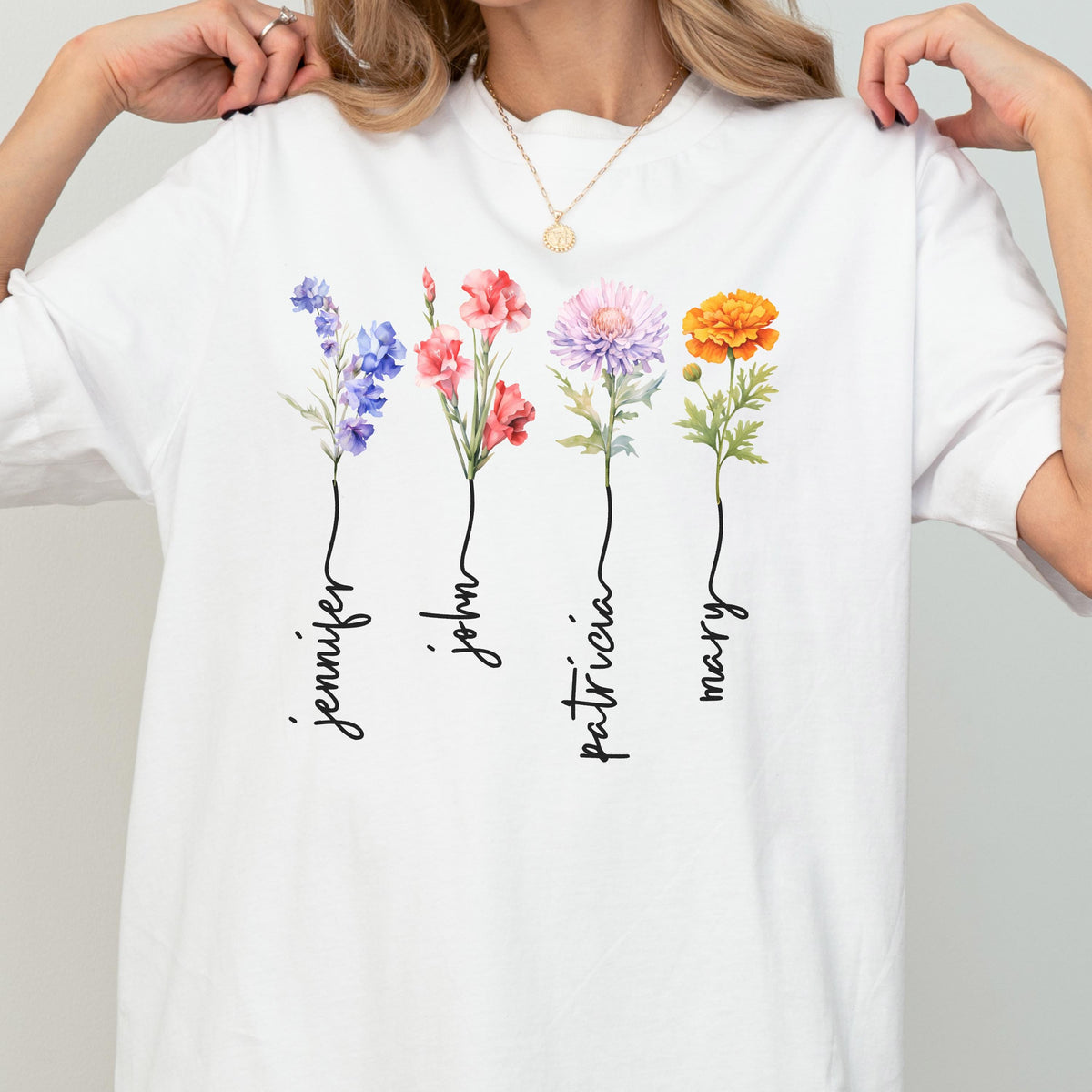 May include: White t-shirt featuring watercolor flowers in shades of purple, red, pink, and orange. Each flower has a name written below it: Jennifer, John, Patricia, and Mary. The shirt has a round neckline.