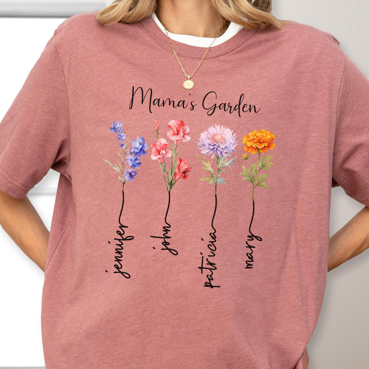 May include: A dusty rose t-shirt with the text "Mama's Garden" and four watercolor flowers. Each flower has a name written below it: Jennifer, John, Patricia, and Mary. The shirt has a white crew neck.