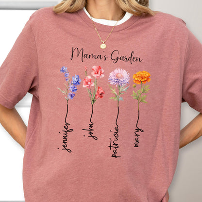 May include: A dusty rose t-shirt with the text "Mama's Garden" and four watercolor flowers. Each flower has a name written below it: Jennifer, John, Patricia, and Mary. The shirt has a white crew neck.