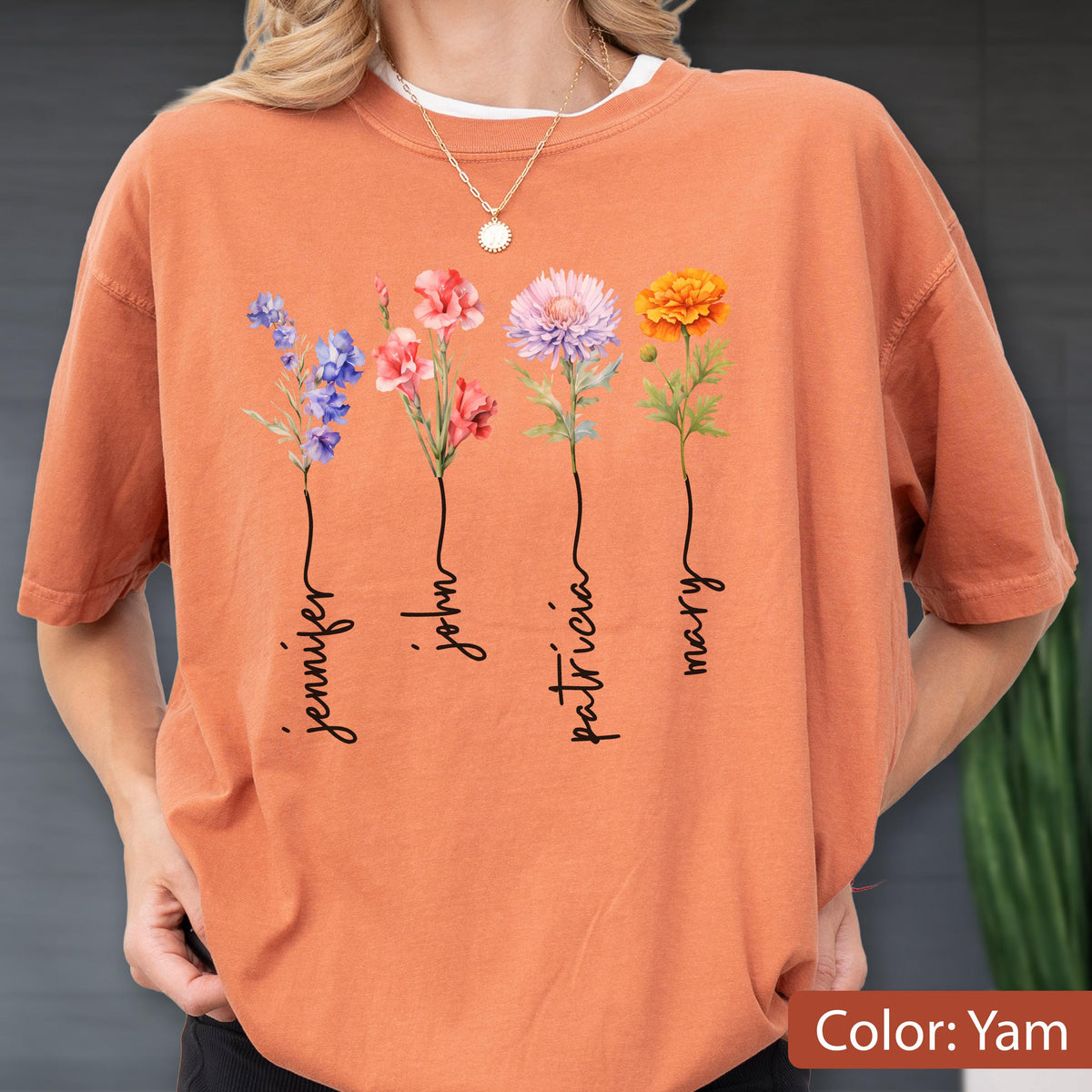 May include: A yam-colored t-shirt featuring four floral stems with names: Jennifer, John, Patricia, and Mary. Each stem has a different flower in shades of blue, pink, purple, and orange. The shirt is a comfortable, casual style.