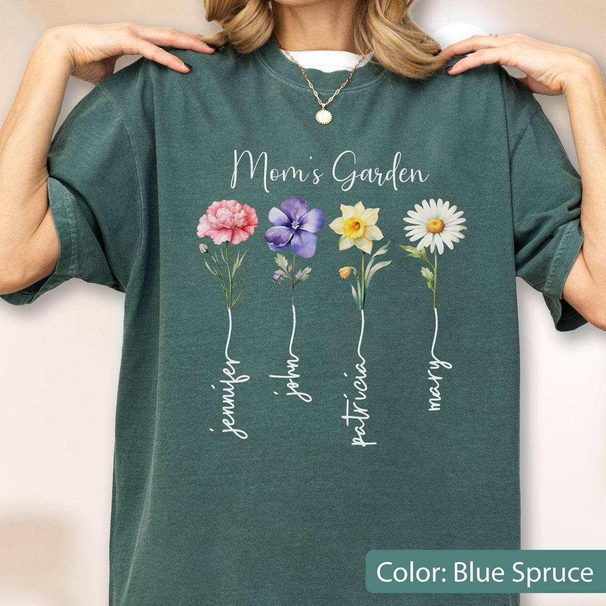 May include: Blue spruce-colored t-shirt with the text "Mom's Garden" and four floral illustrations. Each flower has a name written below it: Jennifer, John, Patricia, and Mary. The shirt has a crew neck.