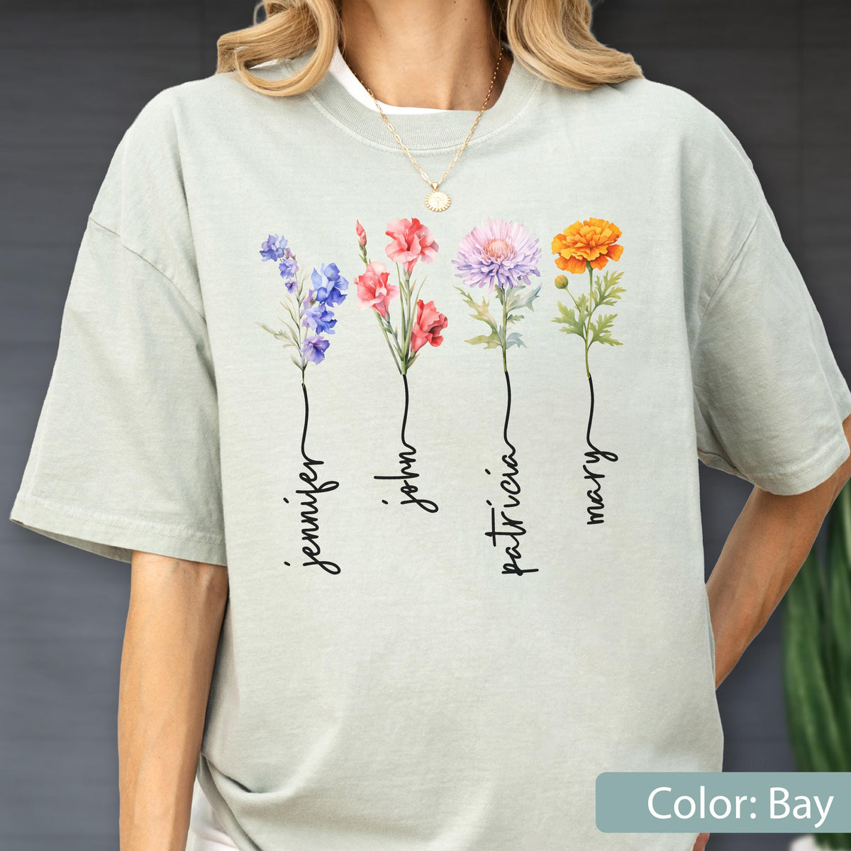 May include: A light green t-shirt featuring four colorful floral illustrations with the names "Jennifer", "John", "Patricia", and "Mary" written below each flower. The shirt is a solid color and has a relaxed fit.