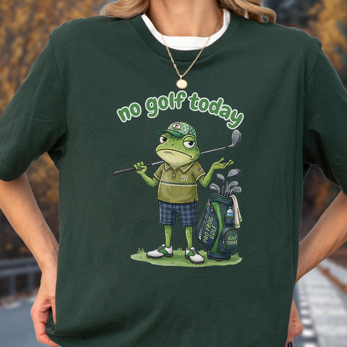 May include: Dark green t-shirt with the phrase "no golf today" in a whimsical font. The shirt features a cartoon frog dressed in golf attire, holding a golf club and standing next to a golf bag. The frog is wearing a golf cap and shorts.