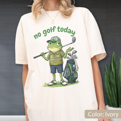 May include: Ivory-colored t-shirt featuring a cartoon frog with a golf club, golf bag, and the text "no golf today." The frog is wearing a golf hat, polo shirt, and plaid shorts. The golf bag has the text "Pro Frog Golf" and "Golf Today."