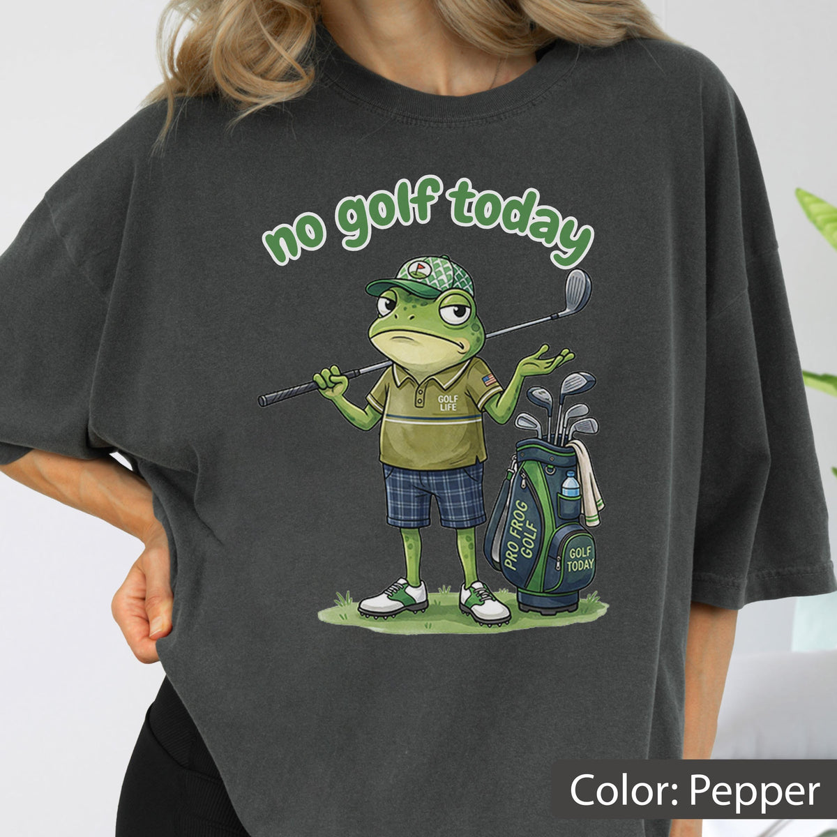 May include: Pepper-colored t-shirt featuring a cartoon frog golfer with the text "no golf today." The frog is wearing a golf hat, polo shirt, and plaid shorts, holding a golf club. A golf bag with clubs is beside him.