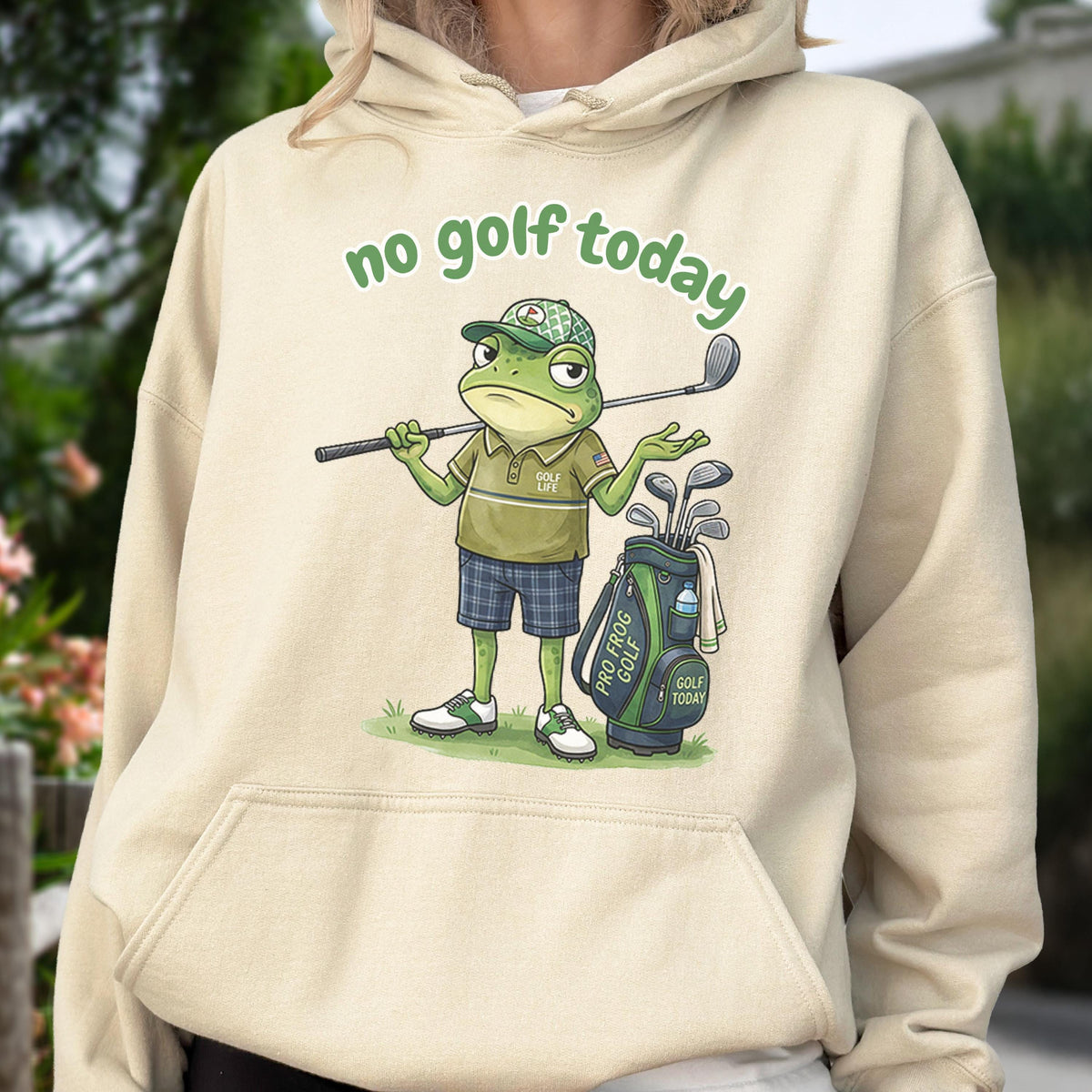 May include: Beige hoodie with a cartoon frog golfer holding a golf club and a golf bag. The frog is wearing a green golf shirt, plaid shorts, and a golf hat. Text on the hoodie says "no golf today". The golf bag has the text "Pro Frog Golf" and "Golf Today".