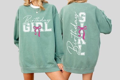 May include: A sage green sweatshirt with the words "Birthday GIRL" in white block letters and a pink bow graphic. The back of the sweatshirt also features the words "Birthday GIRL" in a vertical design with a pink bow.